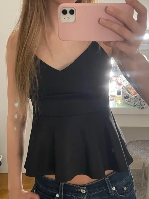 Svart peplum linnetopp med v-ringning - Säljer en svart peplum linnetopp med smala axelband och v-ringad hals. Toppen har en figurnära överdel och en utsvängd nederdel som ger en snygg siluett. 