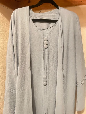 Ljusblå långklänning med knappar - Säljer en ljusblå långklänning med rund halsringning och dekorativa tygklädda knappar framtill. Klänningen har ett mjukt, krinklat tyg och långa, vida ärmar med diskreta sömdetaljer. Enkel och stilren design som ger en chill vibe.