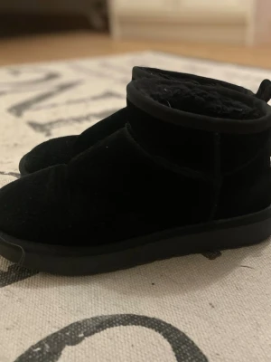Svarta uggs - Säljer ett par svarta uggs med platt sula. Skorna har ett fluffigt foder på insidan som gör dem extra sköna under kyliga dagar. Perfekta att dra på sig när det är kallt ute.