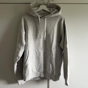 Ljusgrå hoodie med huva och ficka - En stilren ljusgrå hoodie med huva och snörning. Tröjan har en stor magficka och ribbade muddar vid ärmslut och nederkant. Perfekt för dig som gillar en enkel och avslappnad look. Aldrig använd