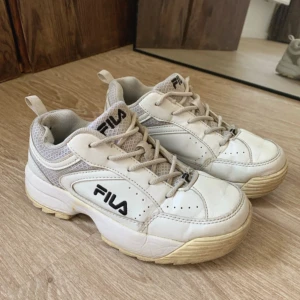 Vita chunky sneakers från Fila - Säljer ett par vita chunky sneakers från Fila med beige detaljer och svart logga på sidan. Skorna har snörning, meshpartier för extra andningsförmåga och en rejäl platt sula. Perfekta för dig som gillar retrostil och vill ha bekväma sneakers till vardags.