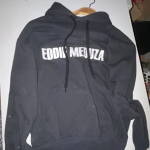 Svart Eddie Meduza hoodie med tryck - Svart hoodie med vit text 'EDDIE MEDUZA' på bröstet och stort tryck på ryggen med Eddie Meduzas ansikte och texten 'RUNKEBALL'. Tröjan har huva med snörning och en stor magficka. Perfekt för dig som gillar kult och vill sticka ut. Använd 2 gånger och de på första bilden är ej smuts utan något som flög i luften och fastnade på kameran.