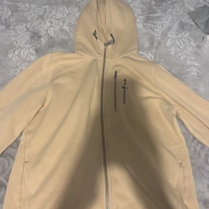 Beige hoodie från Sail Racing med huva - Beige hoodie från Sail Racing med hel dragkedja, huva och bröstficka med dragkedja. Insidan är fodrad med mjuk fleece för extra värme. Perfekt för kyliga dagar och snygg att matcha med jeans eller joggers.