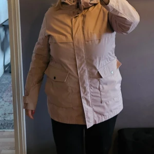 Skön rosa parkasjacka med huva - Snygg ljus rosaparkasjacka med huva och stora fickor framtill. Jackan har långa ärmar, dragkedja och knäppning framtill samt justerbara muddar vid ärmslut samt midja. 