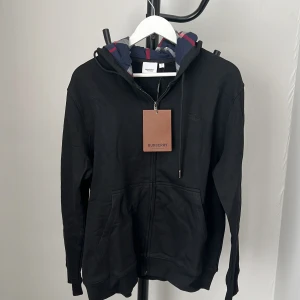 Svart hoodie från Burberry med mönstrad huva - Svart hoodie från Burberry med dragkedja och klassisk logga broderad på bröstet. Huvan har Burberrys ikoniska rutiga mönster i blått, grått och rött. Tröjan har känguruficka och ribbade muddar. Perfekt för dig som gillar stilrena streetwear-plagg.