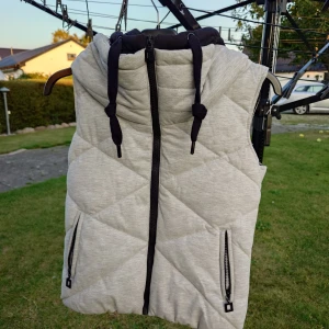 Ljusgrå quiltad dunväst med huva - Snygg ljusgrå dunväst med svart huva och dragkedja framtill. Västen har två fickor med dragkedjor och ett quiltat mönster i diagonala rutor. Perfekt för lager-på-lager och chill stil. Fb sister stl xs