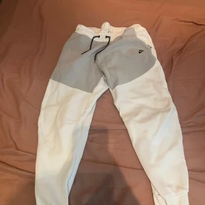 Nike mjukisbyxor i vitt och grått L - Nike mjukisbyxor i storlek L med snygg färgblock-design i vitt och ljusgrått. Byxorna har elastisk midja med svart snörning och en liten Nike-logga på låret. Tillverkade i mjukt bomullsmaterial för maximal komfort.