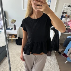 Svart volangblus med kort ärm - Säljer en svart blus med volang nertill och korta ärmar. Toppen har rund halsringning och en lös, luftig passform som ger en avslappnad vibe. Perfekt att styla med jeans eller kjol för en enkel men snygg look.