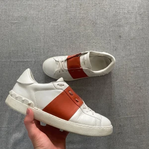 Valentino Garavani Open sneakers vit/röd - Snygga Valentino Garavani Open sneakers i vitt skinn med bred röd läderdetalj över mitten. Klassisk låg modell med snörning och ikoniska nitar på hälen. Perfekta för dig som vill sticka ut med exklusiva och trendiga sneakers. Kan få ned i pris vid snabb affär.