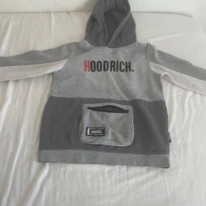 Grå hoodie från Hoodrich med ficka - Säljer en hoodie från Hoodrich med stor magficka med dragkedja och tryckt logga på bröstet. Tröjan har huva, långa ärmar med vita detaljer och är perfekt för streetwear-stil. Mjuk och skön bomullsblandning.