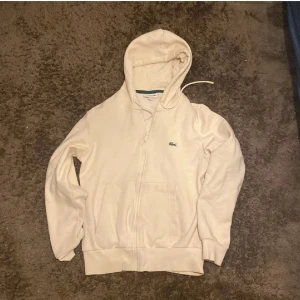 Vit hoodie från Lacoste med dragkedja - Säljer en stilren vit hoodie från Lacoste med klassisk krokodillogga på bröstet. Tröjan har huva med snörning, dragkedja framtill och två fickor. Mjuk insida och ribbade muddar. Perfekt för en chill och clean look.