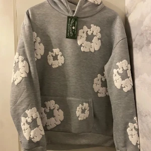 Grå hoodie från Denim Tears med vita detaljer - Grå hoodie från Denim Tears med unika vita blommönster i 3D-effekt över hela tröjan. Klassisk huva, känguruficka och mjukt material som känns riktigt skönt. Perfekt för dig som vill sticka ut med en streetstyle-look.