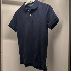 Marinblå pikétröja Polo Ralph Lauren - Klassisk marinblå pikétröja från Polo Ralph Lauren i storlek L (14-16). Tröjan har korta ärmar, krage, två knappar och den ikoniska röda broderade loggan på bröstet. Tillverkad i bomull för en skön och stilren känsla. Inga defekter på tröjan, är lite skrynklig på bilden.