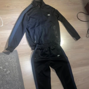 Svart Lacoste Tracksuit - Svart tracksuit från Lacoste med marinblå ränder längs ärmarna och dragkedja framtill. Jackan har två fickor med dragkedja och klassisk Lacoste-logga på bröstet. Perfekt för dig som gillar sportig och stilren look.