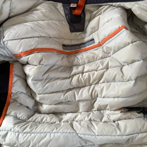 Grå dunjacka från Parajumpers - Säljer en grå dunjacka från Parajumpers med quiltad design och orange detaljer. Jackan har hög krage, dragkedja framtill och två fickor med dragkedja. Klassisk PJS-logga på ärmen och snygga svarta axelpartier. Perfekt för kyliga dagar. Men fickan är tyvärr sönder på högersida