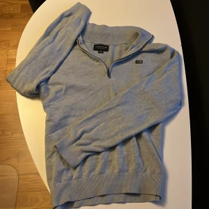 Ljusgrå Lexington half zip tröja - Snygg ljusblå tröja från Lexington med half zip och ribbade muddar. Tröjan har lång ärm och liten Lexington-logga på bröstet. Perfekt för dig som gillar klassisk och stilren look.