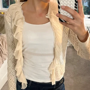 Beige kofta med volangkant - Supersöt beige kofta med volangkant framtill och genombrutet stickat mönster. Långärmad och perfekt att slänga över ett linne eller topp. 