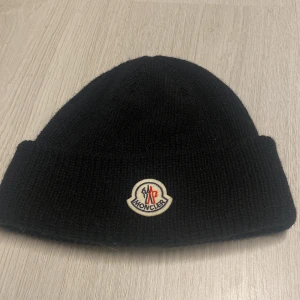 Svart mössa - Svart ribbstickad mössa från Moncler med klassisk uppvikt kant och broderad logotyp framtill. Tillverkad i mjuk ull som håller dig varm under kyliga dagar. Enkel och stilren design som passar perfekt till vinterlooken.