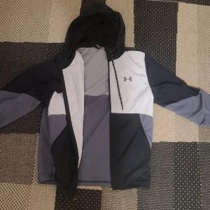 Under Armour vindjacka med huva - Snygg vindjacka från Under Armour i svart, grått och vitt. Jackan har huva med dragsko, dragkedja framtill och sportig design med logga på bröstet. Perfekt för dig som gillar en stilren och funktionell look.