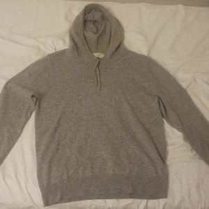 Grå kashmirhoodie från Sayless - Mjuk och lyxig grå hoodie från Sayless i 100% kashmir. Tröjan har huva med snörning och långa ärmar, perfekt för dig som vill ha något riktigt skönt och stilrent. Ribbstickade muddar vid ärmslut och nederkant ger en snygg passform. Ordinarie pris 899