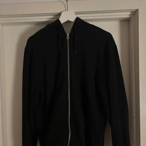 Svart hoodie med dragkedja från Weekday - Svart hoodie från Weekday i storlek S. Tröjan har hel dragkedja framtill, huva med snörning och långa ärmar. Tillverkad i mjuk bomullsmix, perfekt för en chill och stilren look.