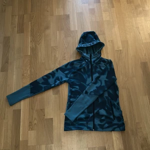 Turkos blå jogg zip Hoodie ifrån peak performance - Säljer en blå hoodie från Peak Performance i vatten, camomönster. Tröjan har huva, dragkedja framtill och är tillverkad i mjuk fleece. Perfekt för kyliga dagar och outdoor-aktiviteter. Snygg och sportig look med långa ärmar och praktiska fickor. Sitter nära och tight på kroppen för och behålla värmen.