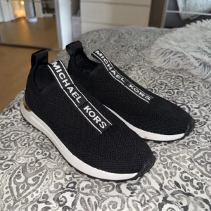 Svarta slip-on sneakers Michael Kors - Svarta slip-on sneakers från Michael Kors med vit sula och gulddetaljer vid hälen. Skorna har stickad ovandel i syntetmaterial och tydlig logga på ovansidan. Perfekta för dig som gillar en sportig och trendig look.