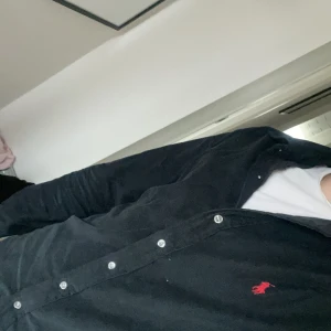 Svart manchesterskjorta Ralph Lauren S - Svart manchesterskjorta från Ralph Lauren i custom fit med klassisk krage och knäppning framtill. Liten röd broderad logga på bröstet. Långärmad modell som passar perfekt till jeans eller chinos. Snygg och tidlös stil med exklusiv känsla.