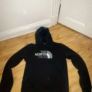 Svart hoodie från The North Face - Svart hoodie från The North Face med stor logga i vitt och grått på bröstet. Klassisk modell med huva, snörning och magficka. Mjuk insida och ribbade muddar vid ärmslut och nederkant. Perfekt för chill eller streetwear.