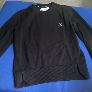 Svart tröja från Calvin Klein - Svart långärmad tröja från Calvin Klein med broderad CK-logga på bröstet och Calvin Klein-text på ärmen. Tröjan har ribbade muddar och nederkant, klassisk rund hals och är tillverkad i mjuk bomull. Perfekt för en stilren och enkel look.