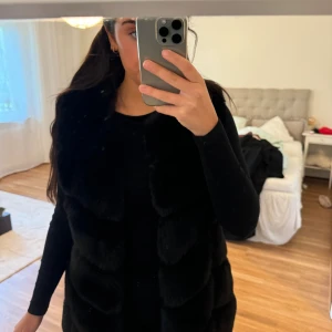 Svart lång pälsväst i fuskpäls - Säljer en svart lång pälsväst i fuskpäls med breda paneler och klassisk krage. Västen är mjuk och fluffig, perfekt för att ge din outfit en lyxig touch. Den har en rak passform och är enkel att matcha med olika stilar.