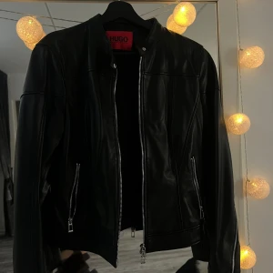 Svart skinnjacka från Hugo Boss - Snygg svart äkta skinnjacka från Hugo Boss med raka linjer och silverfärgade dragkedjor framtill och på fickorna. Jackan har en stilren ståkrage med tryckknapp och är helsvart i färgen. Perfekt för dig som vill ha en klassisk och tidlös look.