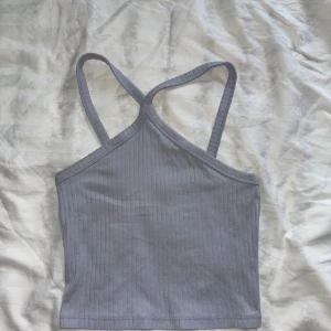 Ljusblå ribbad croppad linnetopp - Säljer en ljusgrå croppad linnetopp med smala axelband som korsas i ryggen. Toppen är ribbad och har en enkel, stilren design som passar perfekt till jeans eller kjol. Materialet är mjukt och stretchigt för en bekväm passform.