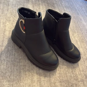 Svarta boots med gulddetalj - Snygga svarta boots med rund tå och grov platt sula. Skorna har dragkedja på sidan och en dekorativ guldfärgad detalj med små stenar och röd accent på utsidan. Perfekta för dig som vill ha en stilren men ändå unik look.