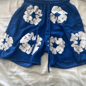 Blå shorts från Denim Tears med tryck - Säljer ett par blå shorts från Denim Tears i mjuk bomull med vita blommiga tryck över hela plagget. De har elastisk midja med snörning och fickor både fram och bak. Perfekta för en chill och trendig sommarstil.