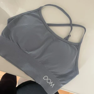 Grå sporttopp från Drop of Mindfulness - Säljer en grå sporttopp från Drop of Mindfulness med smala, justerbara axelband som korsas i ryggen. Toppen har bred ribbad kant nedtill och logga både fram och bak. Perfekt för träning och ger en snygg, sportig look.