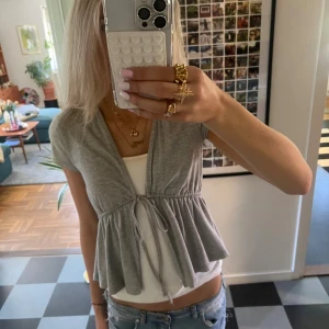 Grå omlottopp med knyt från Gina Tricot - Supersöt grå omlottopp från Gina Tricot med korta ärmar och knyt i midjan. Toppen har v-ringning och volangliknande nederdel. Storlek s💓💋