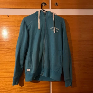 Blå hoodie från Race Marine - Blå hoodie från Race Marine med dragkedja framtill och vita snören i huvan. Tröjan har två fickor på sidorna och tryckt logga på bröstet. Perfekt för chill dagar eller när du vill ha en avslappnad stil. Färgen gör sig inte rättvis på bild. 