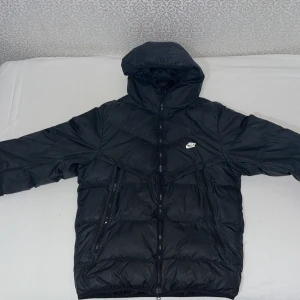Svart pufferjacka från Nike Storm-FIT - Svart pufferjacka från Nike med Storm-FIT-teknologi. Jackan har huva, dragkedja framtill och två stora fickor med dragkedja. Klassisk Nike-logga på bröstet. Perfekt för kalla dagar när du vill hålla stilen och värmen.