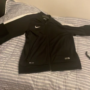 Svart Nike Dri-Fit träningsjacka L - Svart träningsjacka från Nike med vit logga på bröstet och vit detalj längs ena ärmen. Jackan har hel dragkedja framtill och är tillverkad i Dri-Fit polyester som andas. Perfekt för träning eller sportiga outfits.