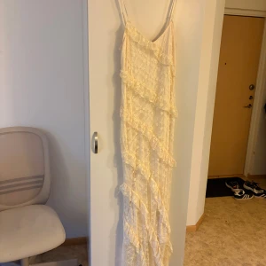 Lång spetsklänning med volanger - Romantisk långklänning i ljusbeige spets med smala axelband och v-ringning. Klänningen har flera lager volanger och ett vackert blommigt spetsmönster över hela tyget. Perfekt för dig som vill ha en dreamy och feminin look. Storlek L men sitter som M