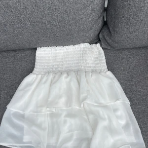 Vit volangkjol  - Supersöt vit kortkjol med bred smock i midjan och dubbla volanger. Kjolen är gjord i ett tunt, lätt och glansigt material som ger en luftig känsla. Perfekt för dig som gillar en feminin och trendig stil.