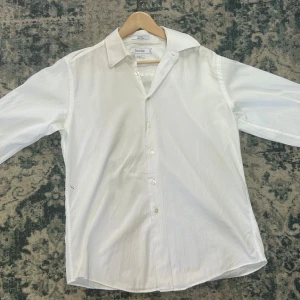 Vit slim fit skjorta från Calvin Klein - 34/35. Snygg vit skjorta från Calvin Klein i slim fit-modell. Skjortan är tillverkad i 100% bomull och har diskreta ränder i tyget, klassisk krage och knäppning framtill. Perfekt för dig som gillar stilrena och fräscha plagg.  Det finns en liten fläck på sidan men den går lätt att tvätta bort