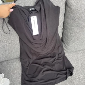 Svart kortklänning från Gina Tricot - Svart kortklänning från Gina Tricot med snygg draperad halsringning. Klänningen är ärmlös och har en stilren, enkel design som känns både trendig och klassisk. Perfekt för dig som gillar minimalistisk stil och vill ha något som sticker ut utan att vara för mycket.