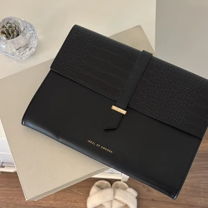 Svart datorväska Ideal of Sweden - Stilren svart datorväska från Ideal of Sweden med gulddetaljer. Perfekt för att bära din laptop med stil. Går ej att köpa längre, köpt för 899, fint skick och endast små repor vid gulddetaljen i fram men inget man tänker på💛