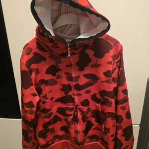 Röd camo hoodie  - Säljer en fet röd camo hoodie med hel dragkedja som går upp över huvan. Huvan har ikoniskt hajmotiv, broderade bokstäver och tigertryck på sidan. Hoodie med känguruficka och cool streetwear-vibe. Perfekt för dig som vill sticka ut. Står storlek S i men passar XS 