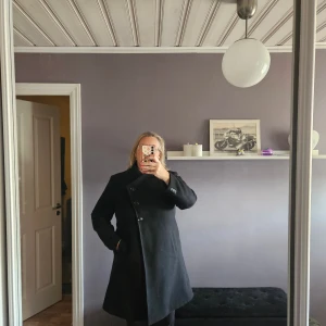 Svart kappa med asymmetrisk knäppning - Stilren svart kappa med snygg asymmetrisk knäppning framtill och klassisk krage. Långärmad modell med fickor på sidorna. Perfekt för dig som vill ha en tidlös och clean look under höst och vinter. Materialet känns som ullblandning.