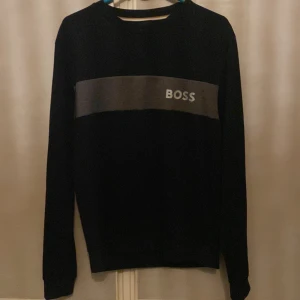 Svart långärmad tröja från BOSS - Svart långärmad tröja från BOSS med en grå panel över bröstet och vit BOSS-logga. Klassisk rund halsringning och normal passform. Tillverkad i mjuk bomull, perfekt för en stilren och enkel look.