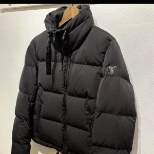 Svart pufferjacka Rockandblue - Svart pufferjacka från Rockandblue med hög krage, dragkedja framtill och snörning i nederkant. Jackan har två sidofickor med dragkedja och en snygg logotyp på ärmen. Perfekt för kalla dagar och har en modern, croppad passform. Nyskick, använd ett fåtal gånger, kvitto finns, nypris 2500-3000.