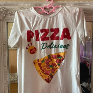 T-shirt No°1 - Vit t-shirt med färgglatt tryck av en pizzaslice och texten 'PIZZA Delicious' i rött och grönt. T-shirten har rund hals och korta ärmar, perfekt för dig som gillar roliga och unika motiv. Materialet känns som bomull och passformen är normal.
